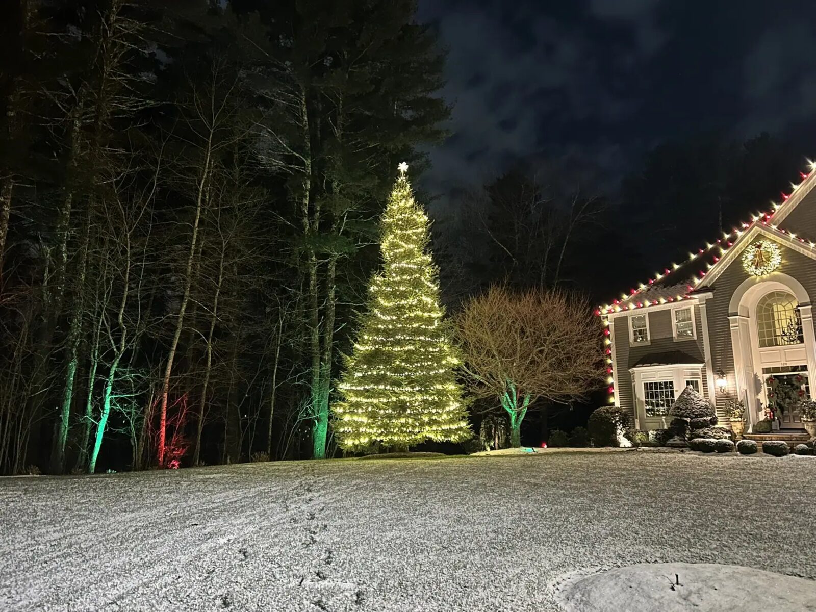 A lit Christmas tree beside a home with warm roofline lighting on a winter evening
